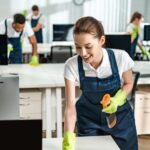 cheerful cleaner in overalls cleaning office desk with rag