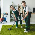 happy young janitors claning carpet and smiling at camera in office
