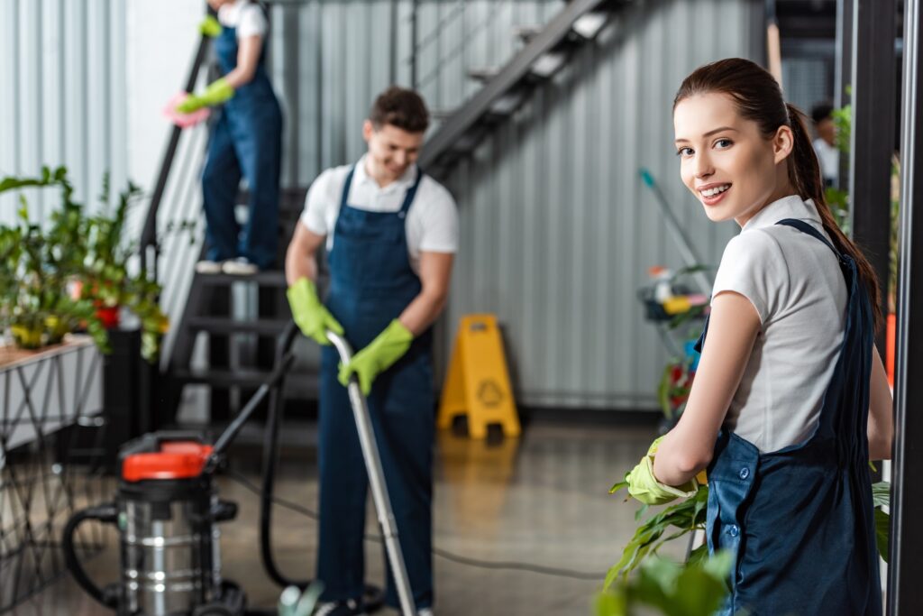 attractive cleaner smiling at camera while colleagues cleaning office