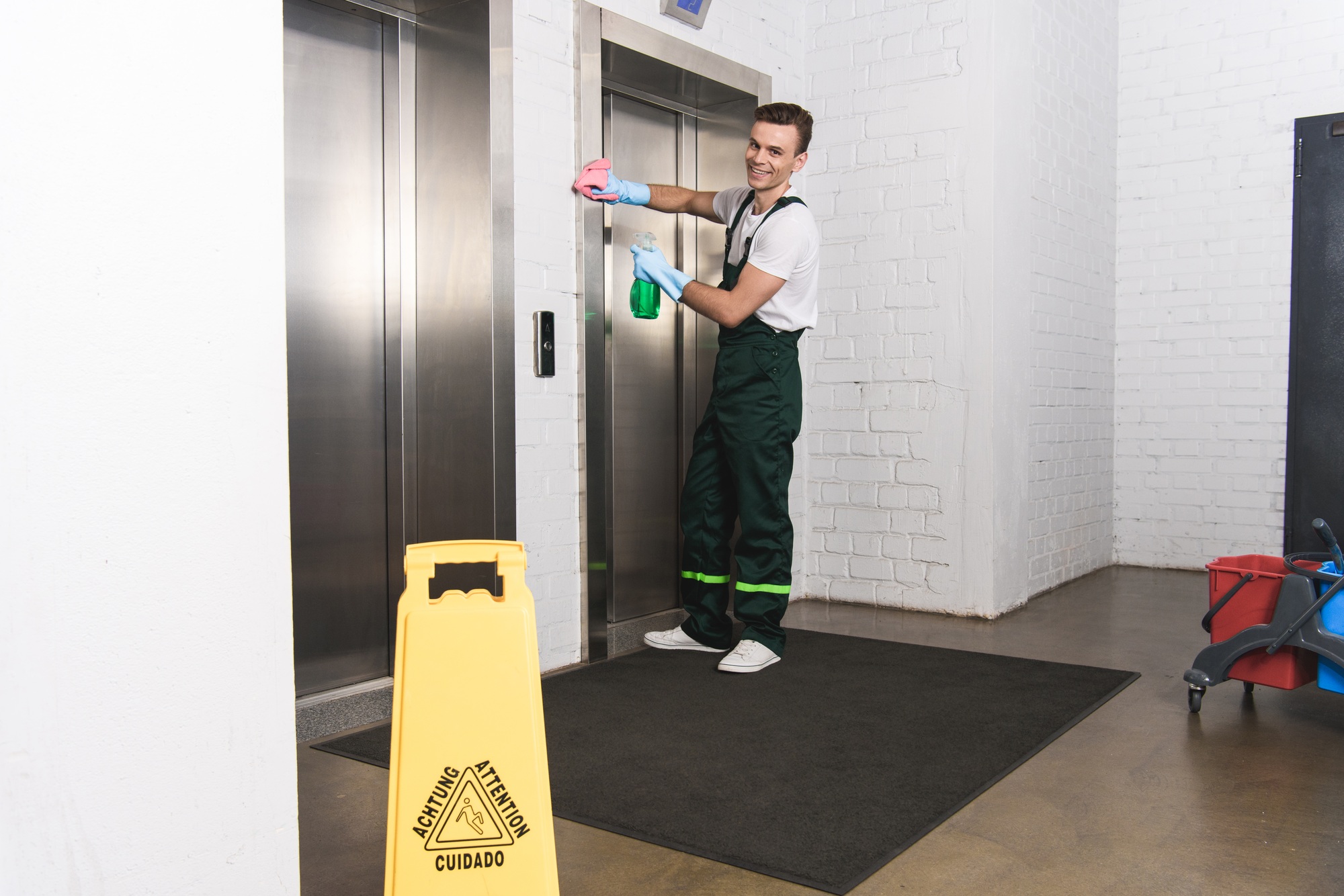 handsome young janitor cleaning elevator and smiling at camera